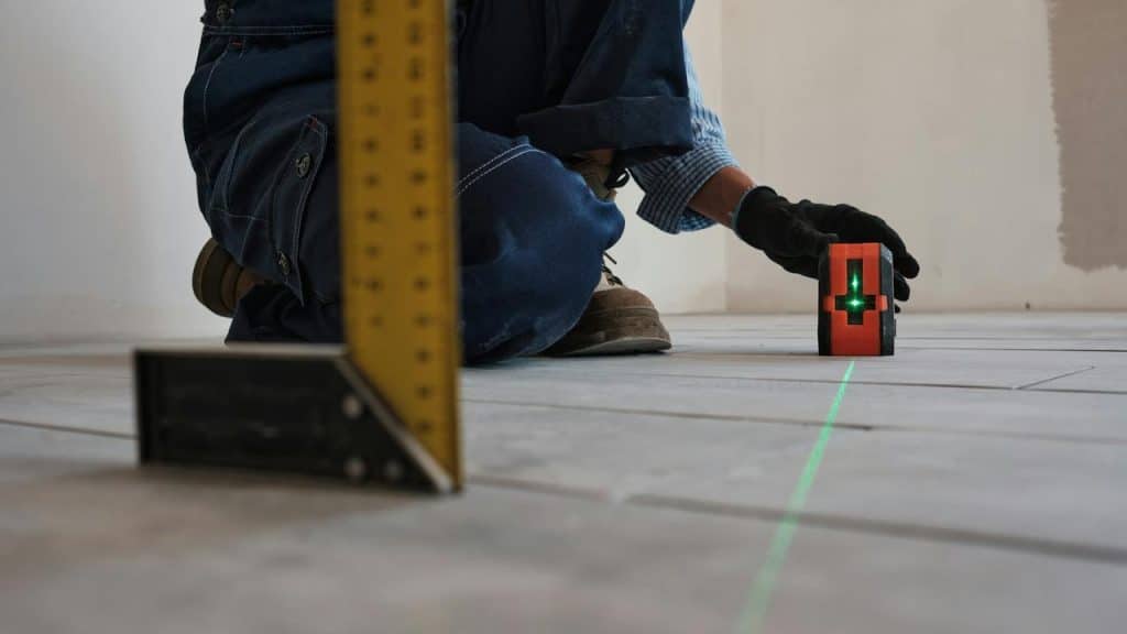 A red laser level projecting green cross lines on a tiled floor beside a right-angle square.