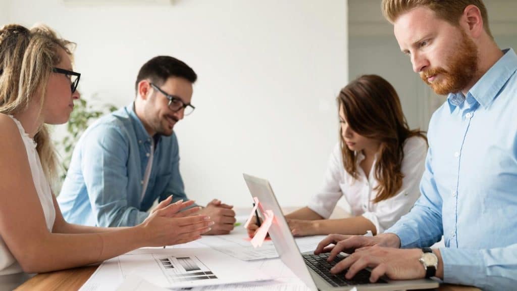 An architect reviewing blueprints and typing on a laptop at a meeting table with colleagues.