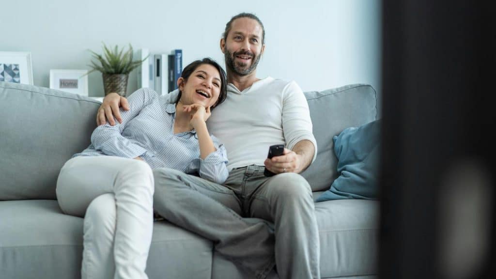 A couple cuddled on a couch watching television in a bright living room.