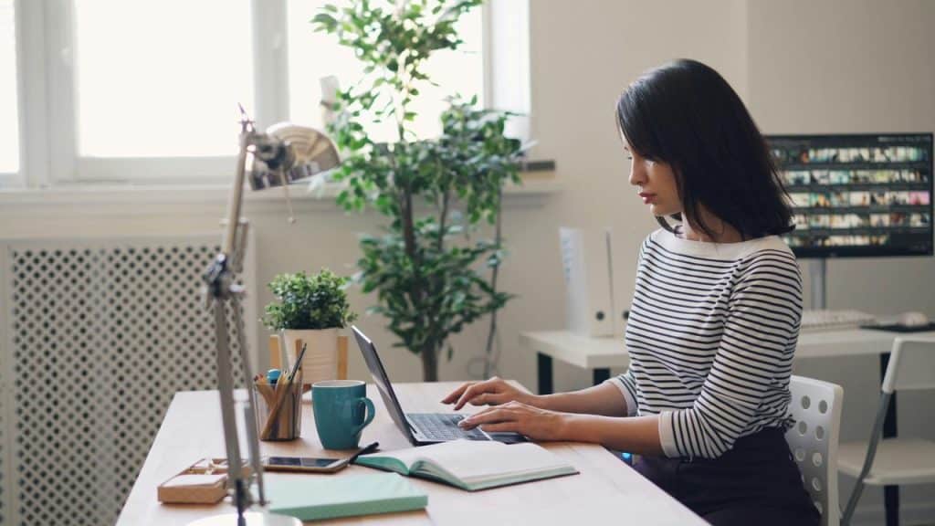 A woman typing on a laptop at a desk in a sunlit home office.