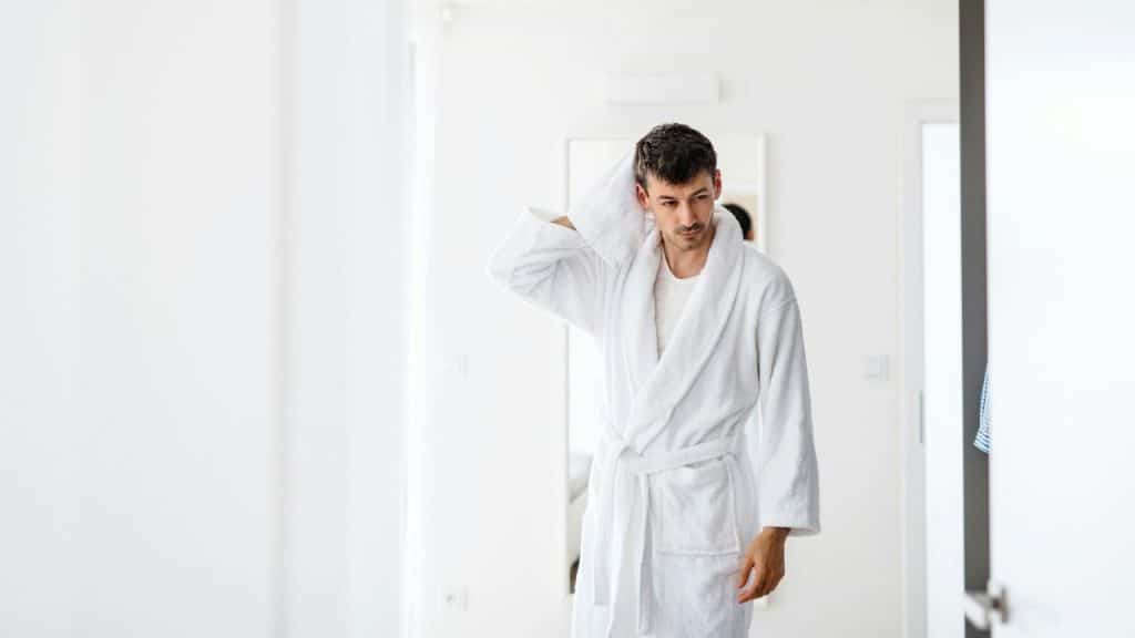 A man in a white bathrobe stands in a brightly lit room.