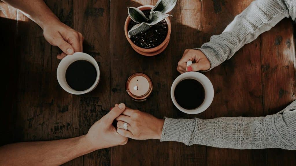 A couple holding hands across a wooden table with two coffee mugs.