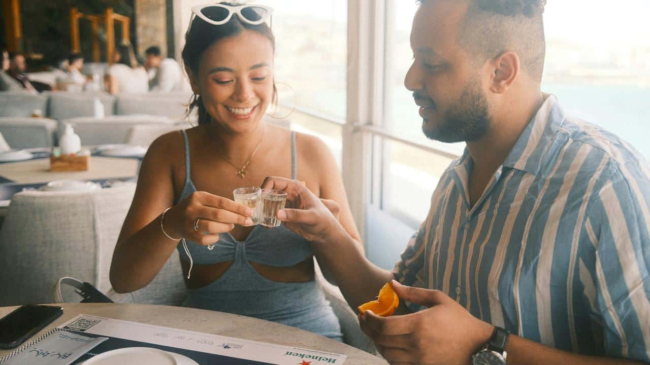 A couple clinking shot glasses at a restaurant table.