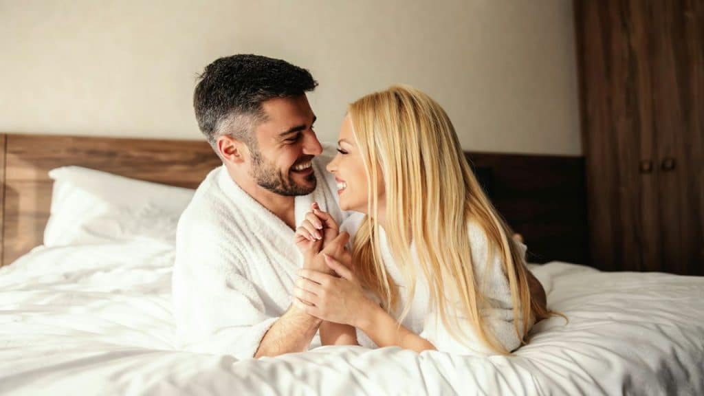 A smiling man and woman in white robes lie on a bed, holding hands and looking at each other.