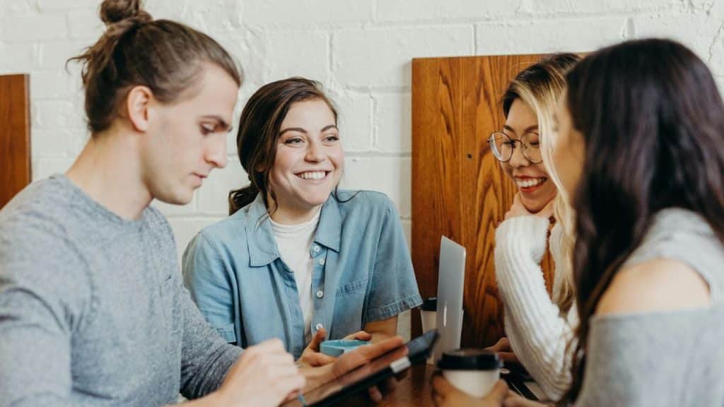 A diverse group of four young adults is happily gathered around a table in a cafe, talking and smiling.