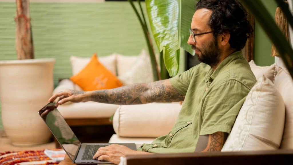 A bearded man with tattoos on his arms uses a laptop on a couch.
