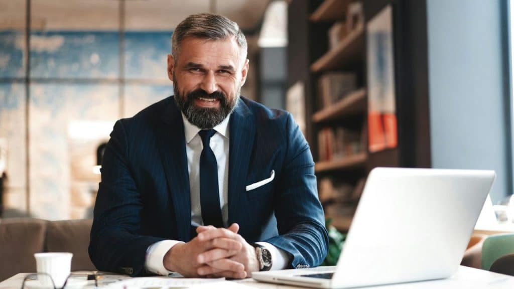 A smiling, bearded man in a dark pinstripe suit sits at a desk with a laptop.