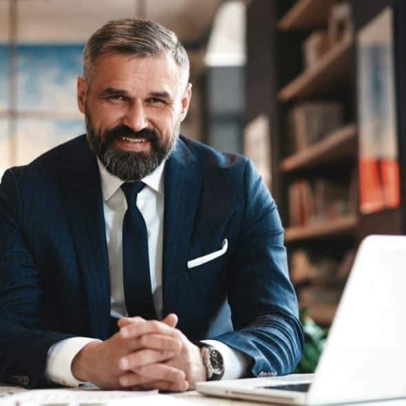 A smiling, bearded man in a dark pinstripe suit sits at a desk with a laptop.
