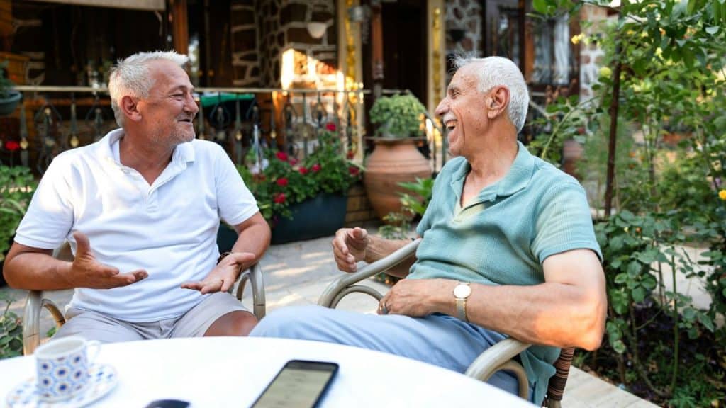 Two senior men are sitting at a table outdoors and laughing.