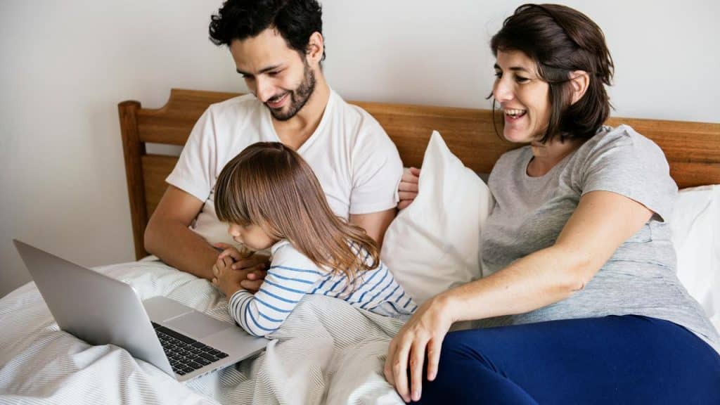 A family of three watching a laptop together in bed.