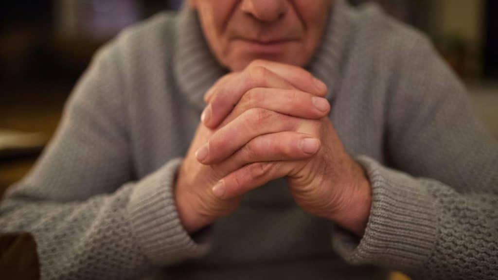 An elderly man’s clasped hands resting on a table.