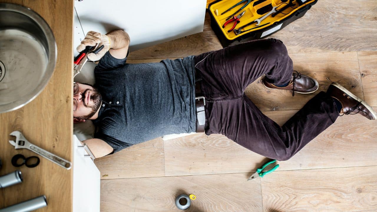 A man lies on a wooden floor, reaching under a sink with tools, fixing a plumbing issue.