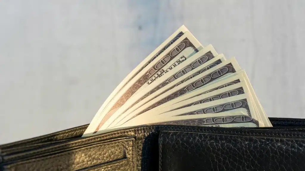 A black leather wallet holding a fanned stack of $100 bills.