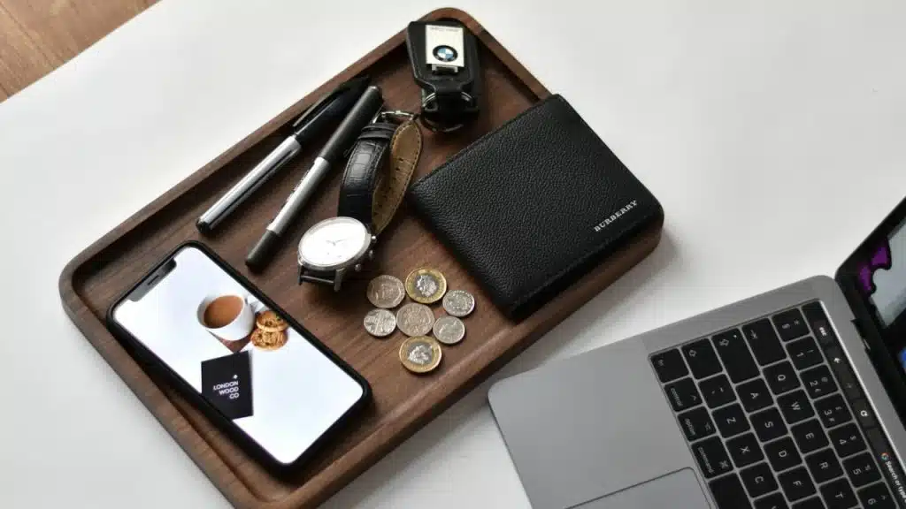 A tray holds a phone, wallet, watch, pens, keys, and coins beside a laptop.