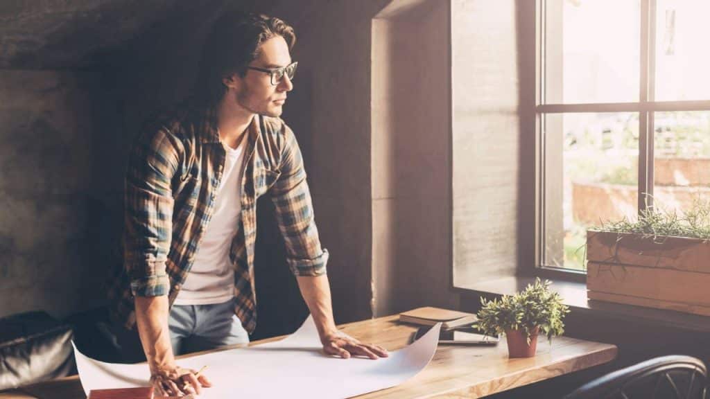 A man in a plaid shirt and glasses leans over papers on a wooden table, looking out a sunlit window.