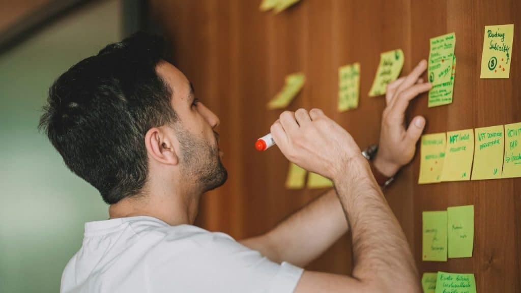 A man in a white shirt writes on a yellow sticky note on a wooden board.