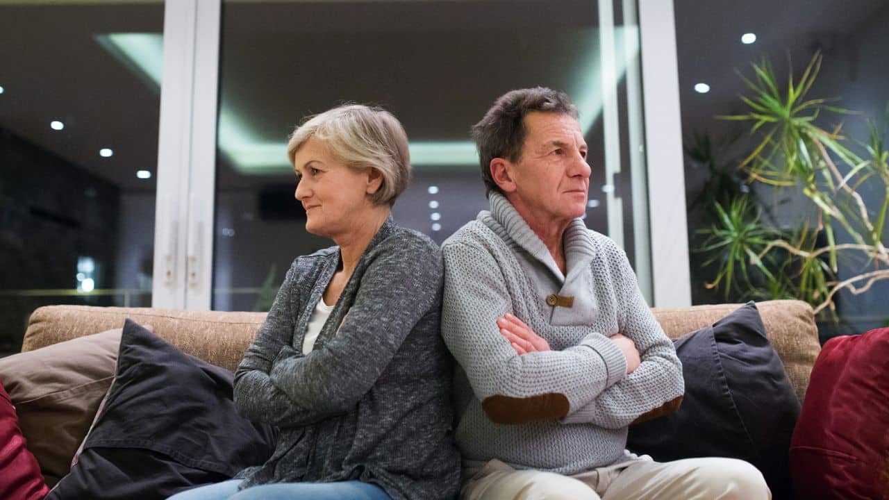 A displeased senior couple sits back-to-back on a couch, arms crossed.