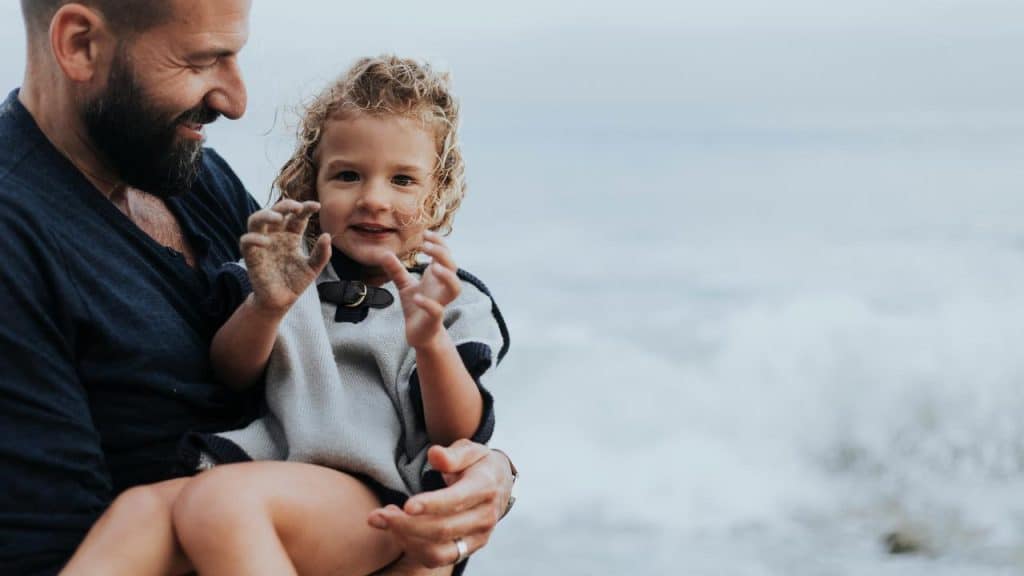 A man holding a sandy-handed child by the sea.