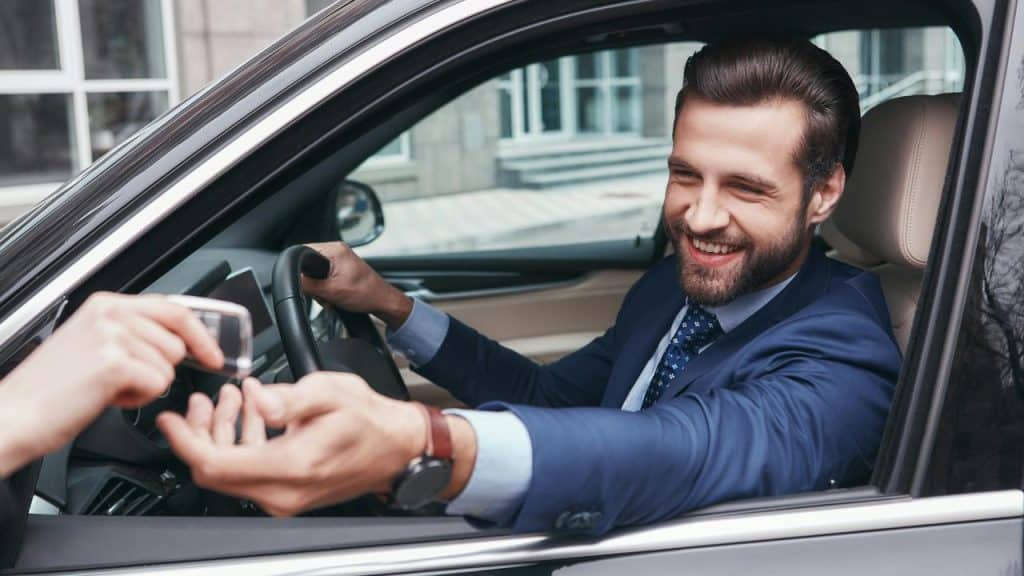 A man in a suit smiling and handing car keys through his car window.