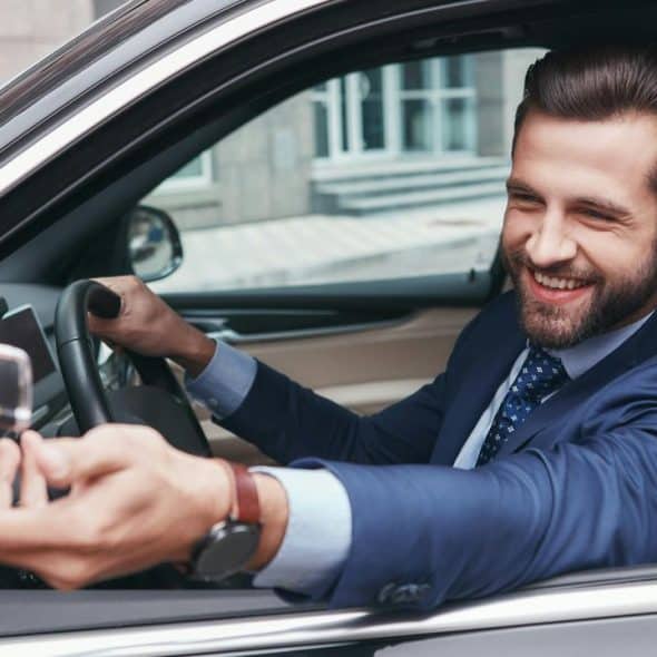 A man in a suit smiling and handing car keys through his car window.