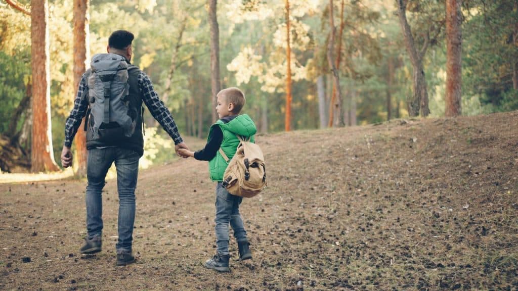 A man with a backpack walking hand in hand with a boy through a forest.