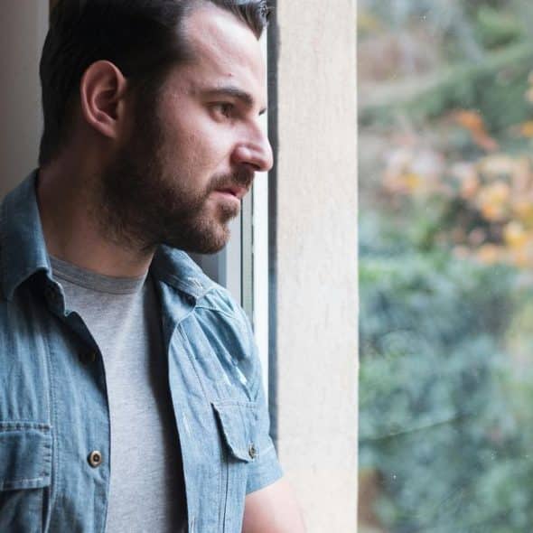 A bearded man looking pensively out a window.