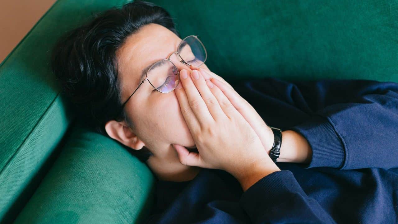 A person wearing glasses lies on a green couch, hands covering their face.