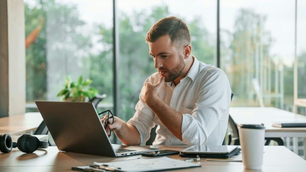 A man in a white shirt looks at his laptop, holding glasses, at a desk.