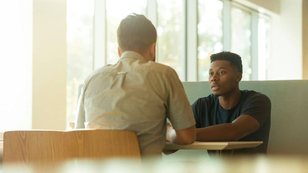 A Black man looks intently at a man with his back to the camera in a bright room.