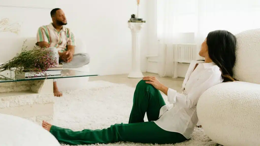 A woman in green pants and a white shirt leans against a sofa, while a man sits on another sofa in the background.