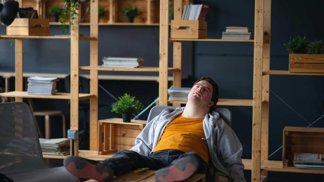 A man slumped back in a chair with eyes closed in an office setting.