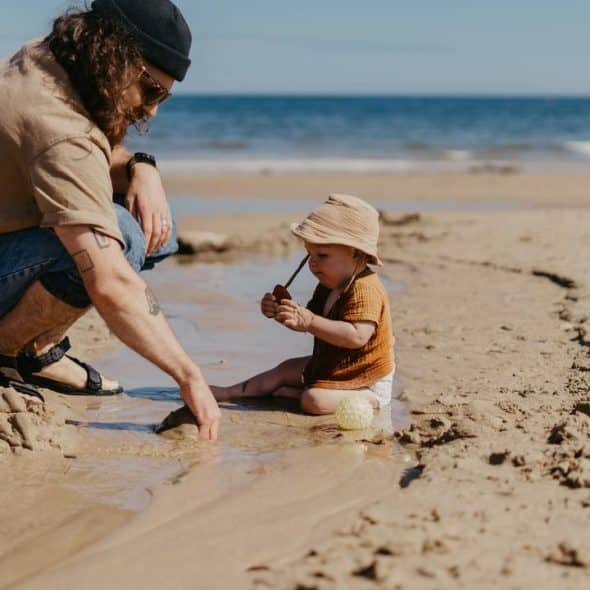 A man crouching beside a toddler playing with sand and water on a beach.