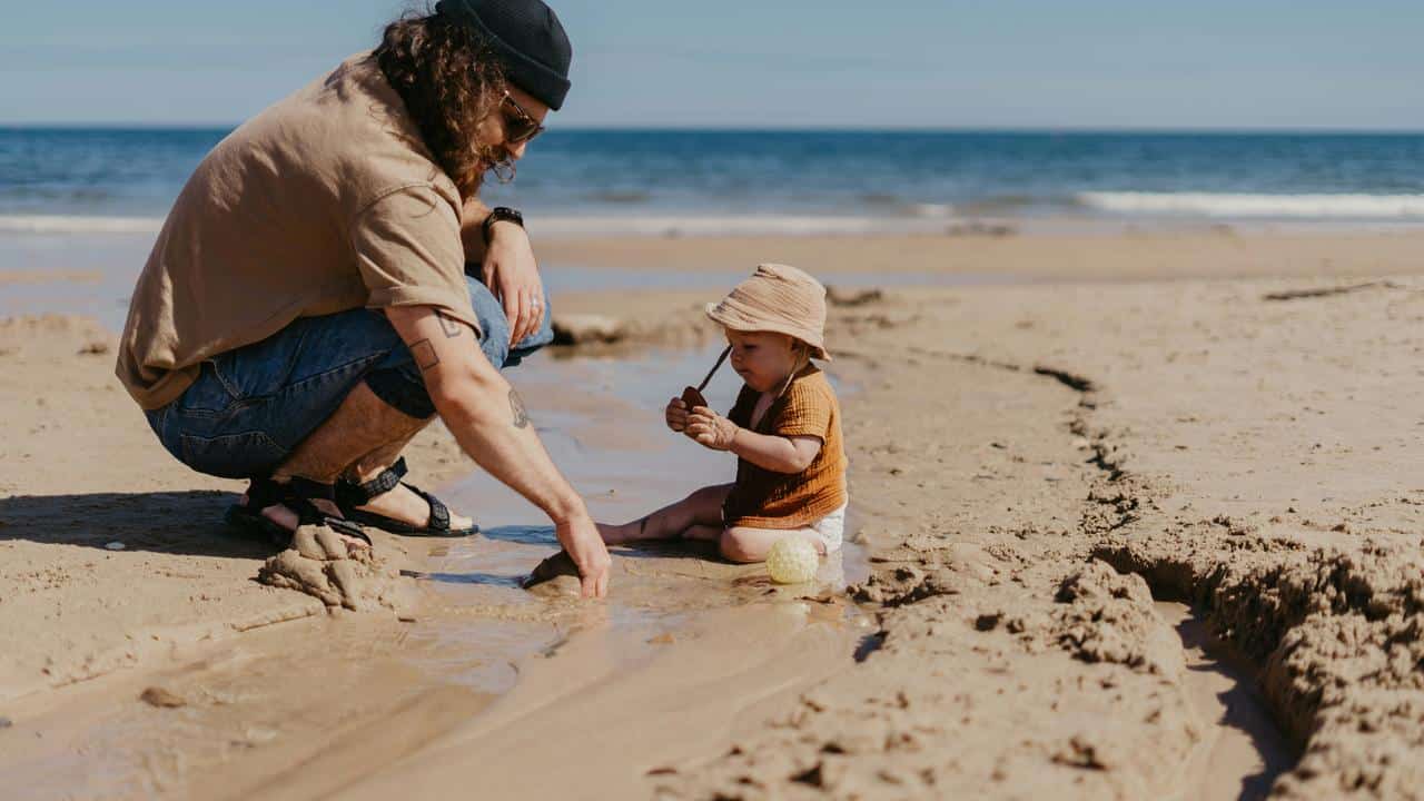A man crouching beside a toddler playing with sand and water on a beach.