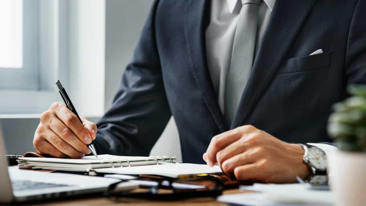 A man in a suit writing in a notebook at a desk.