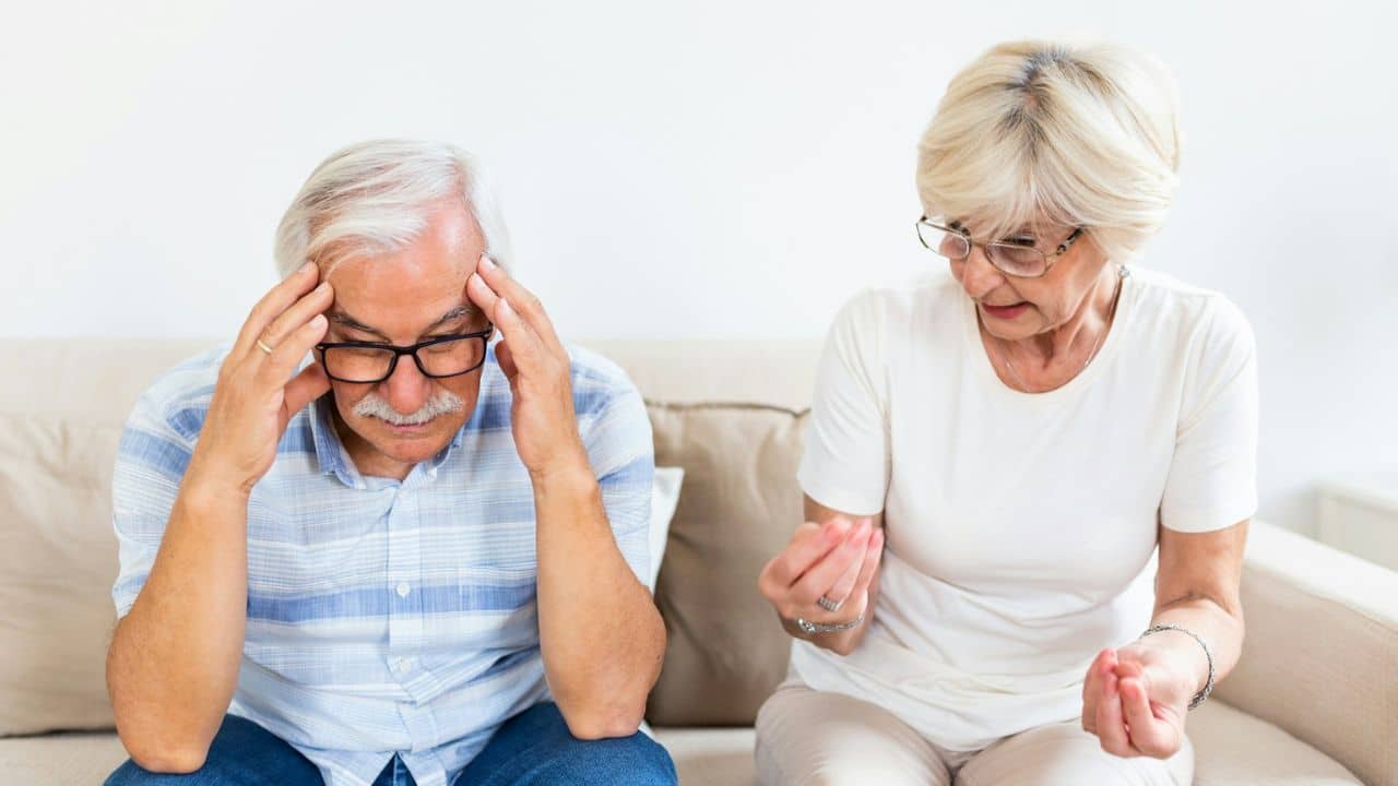 A distressed older man holds his head while an older woman talks to him.