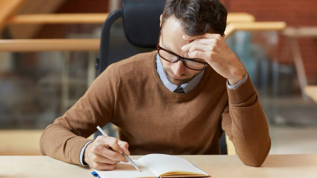A man with glasses, hand on forehead, writes in a notebook at a desk.