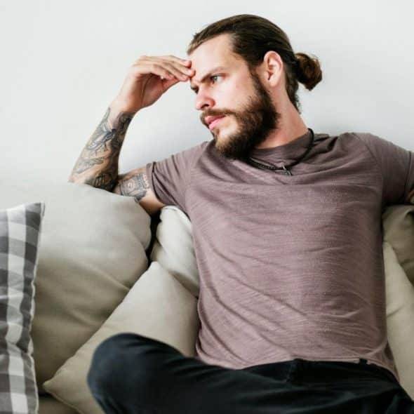 A man with a beard, man bun, and tattooed arms sits on a couch, looking to his right with a pensive expression.
