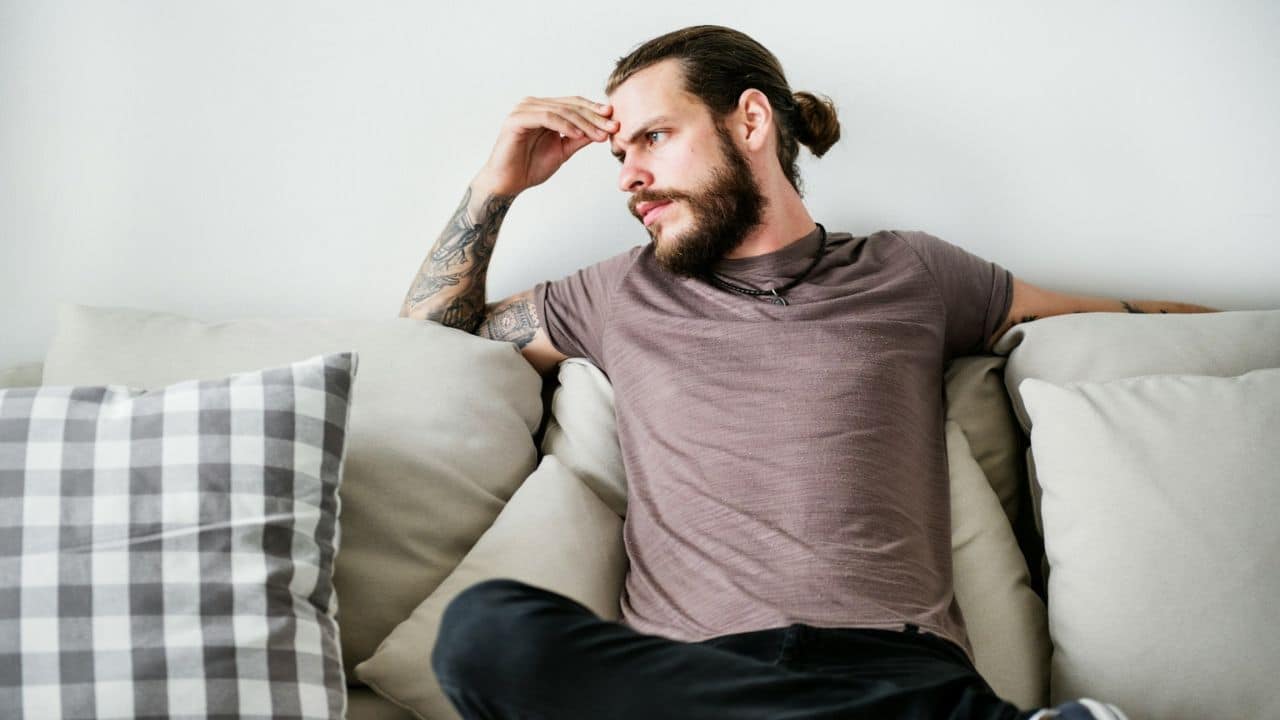 A man with a beard, man bun, and tattooed arms sits on a couch, looking to his right with a pensive expression.