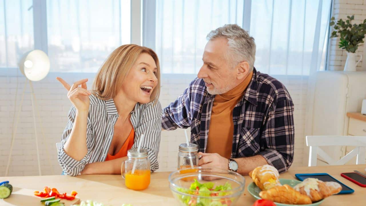 A cheerful couple shares a meal at a table, looking at each other and smiling.