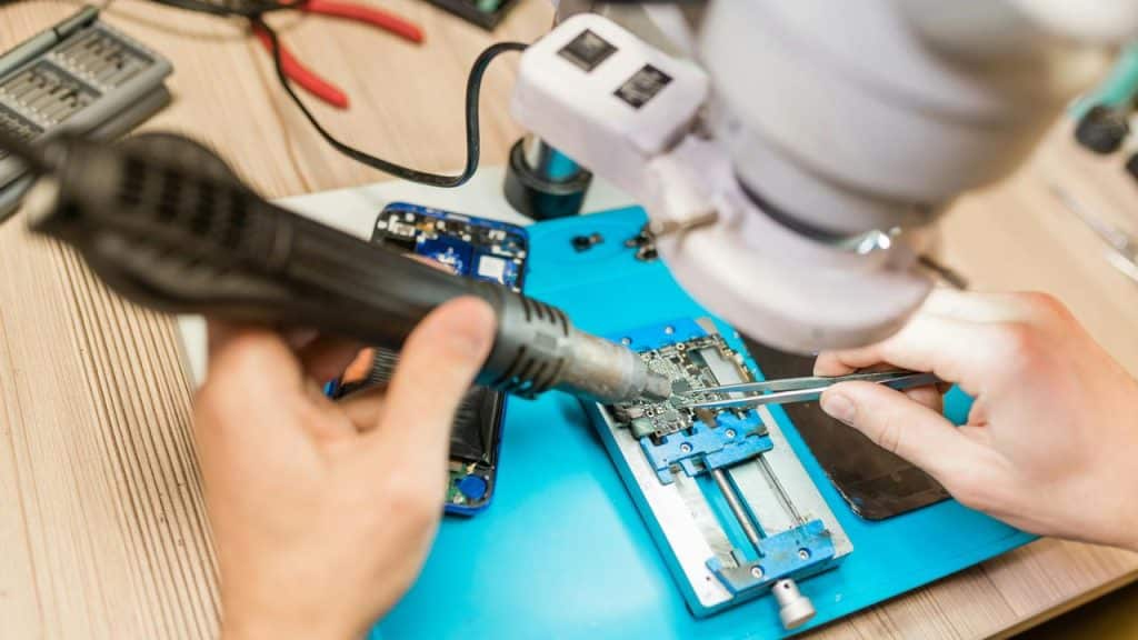 A technician using a heat gun and tweezers to repair a smartphone circuit board.