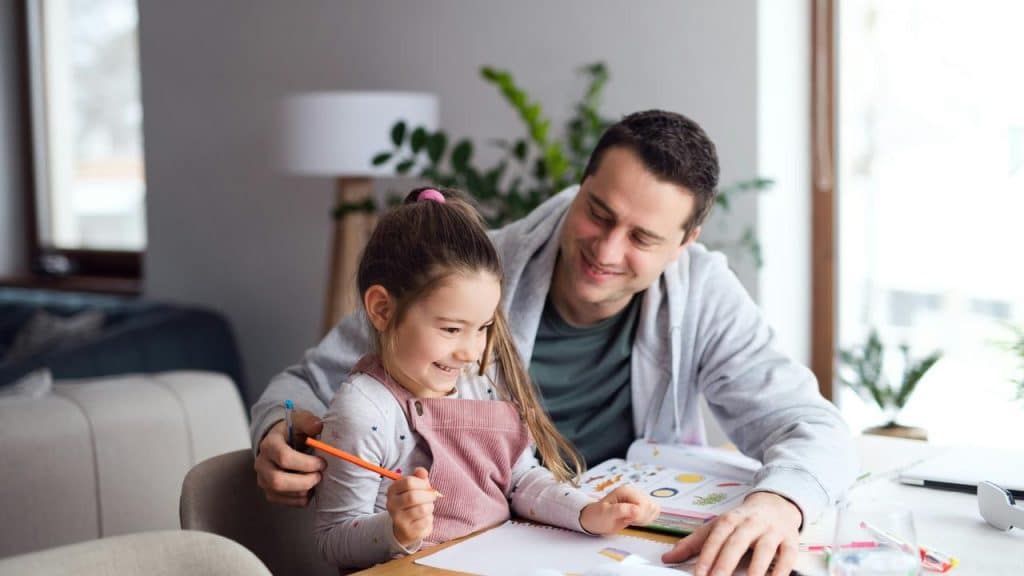 A father guiding his daughter as she draws at a table.