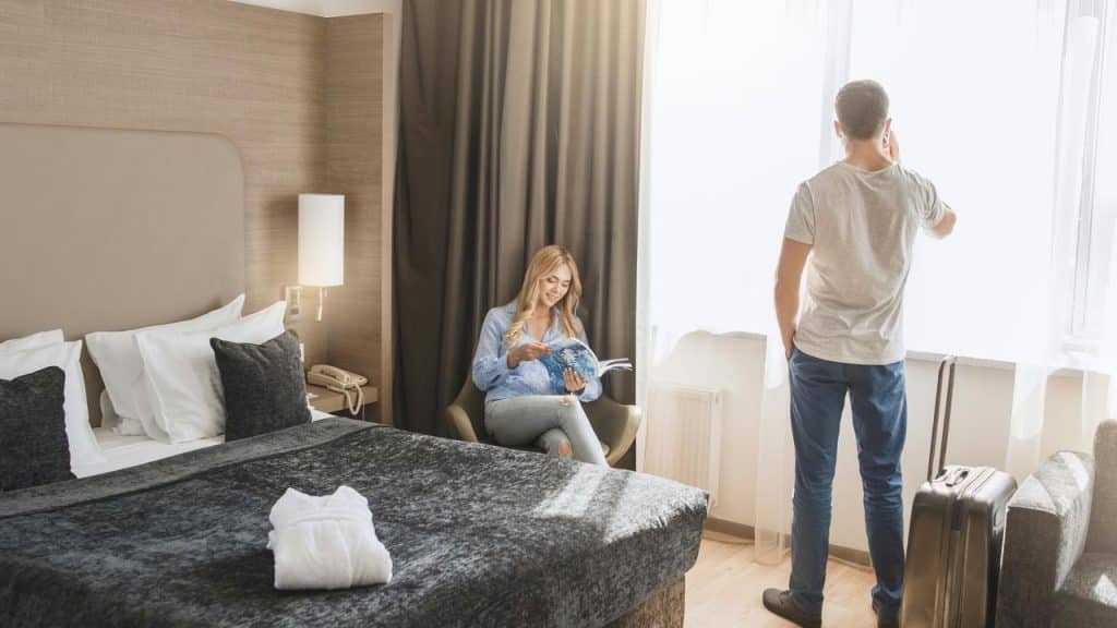 A woman reading a magazine in a hotel room as her partner stands by the window on the phone.