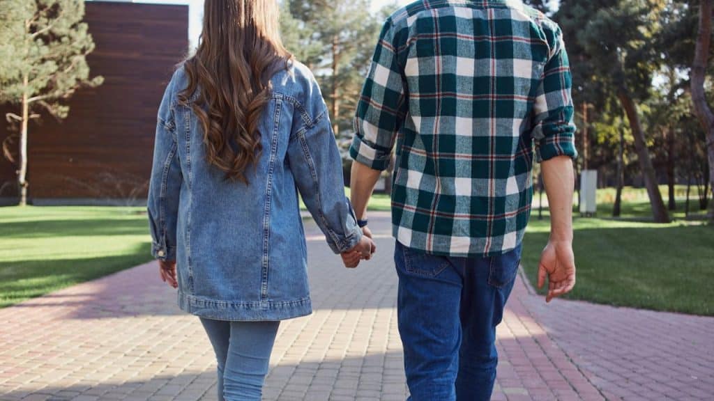 A man and a woman, viewed from behind, hold hands while walking on a paved path.