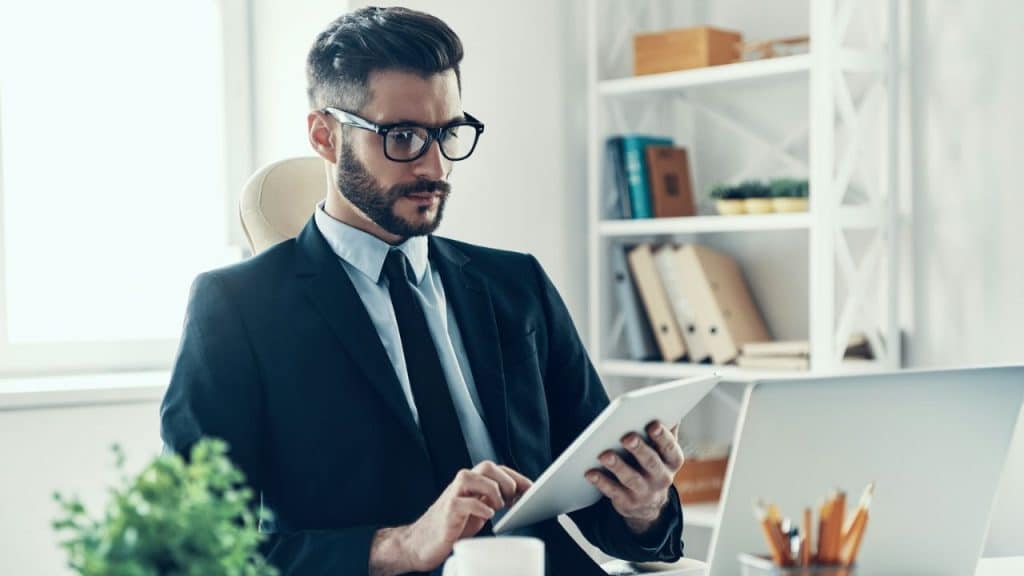 A businessman with glasses and a beard sits at a desk, looking at a tablet.