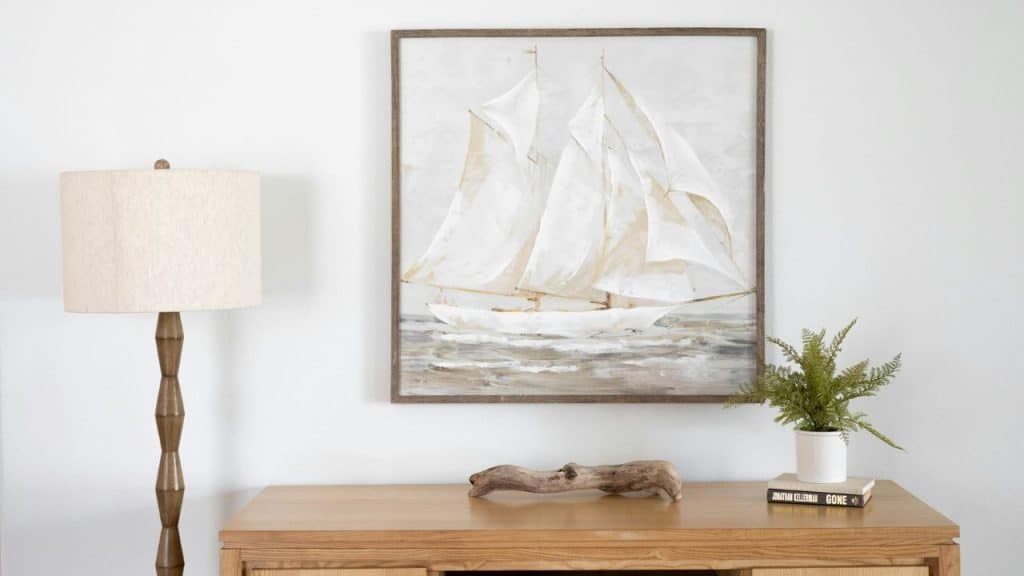A framed sailboat painting hangs above a wooden console table with a lamp and plant.