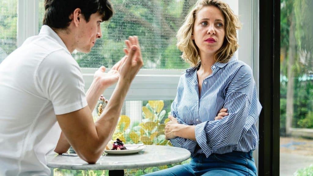 A man talking to an upset woman sitting with arms crossed at a cafe table.