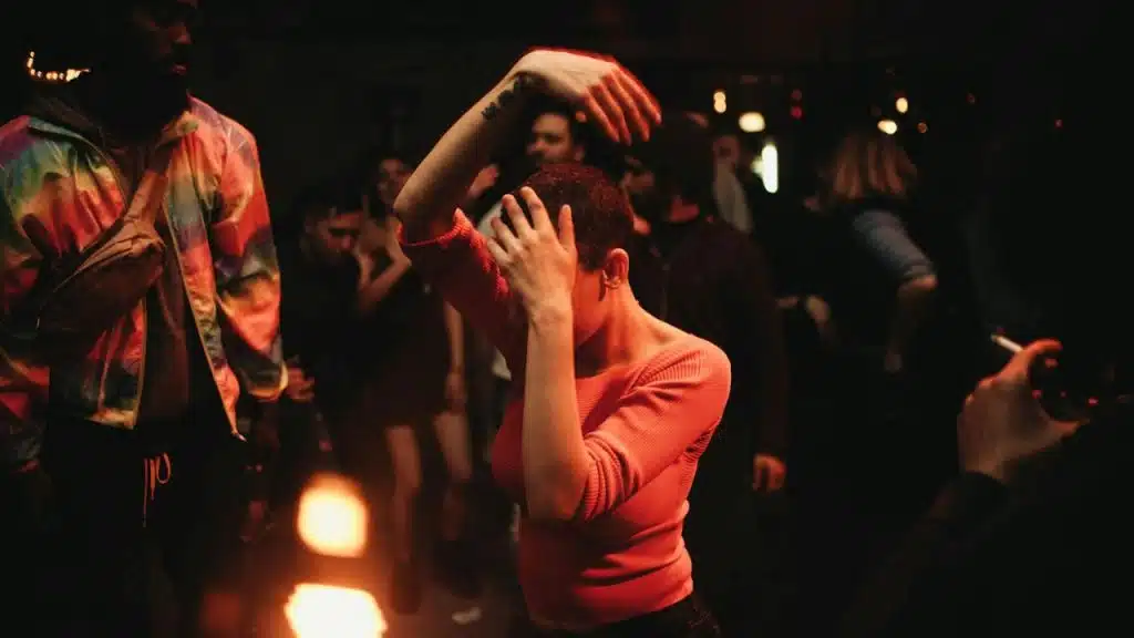 A woman in a pink top with short hair covers her face with her hands, dancing in a dim, crowded club.