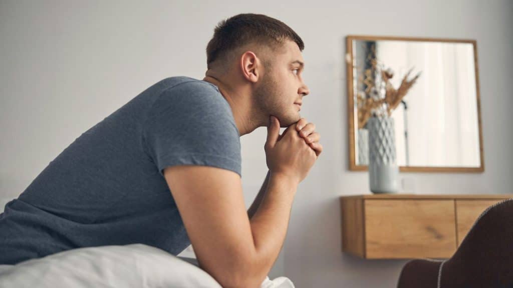 A man in a blue t-shirt sits on a bed, looking thoughtfully to the right.