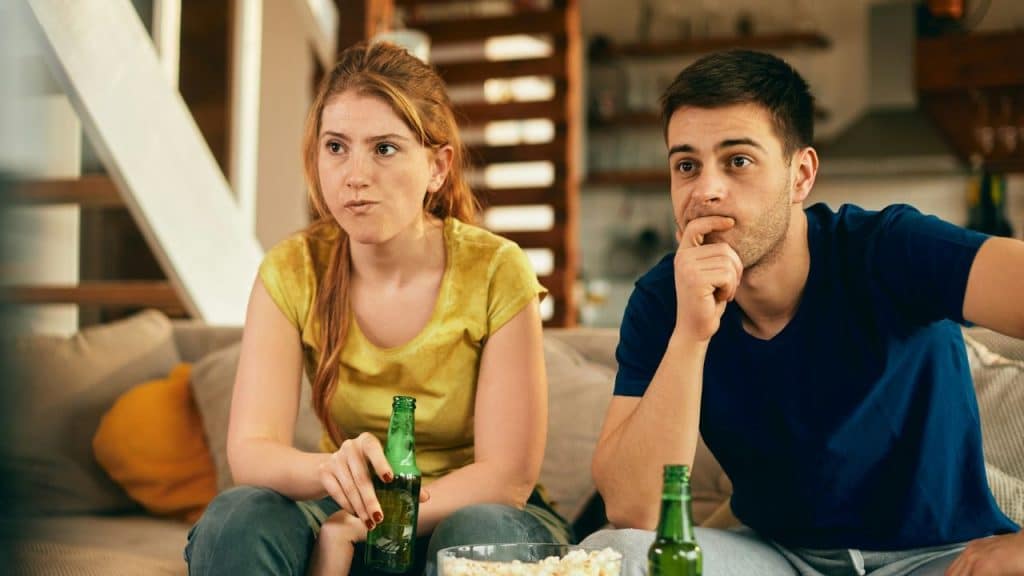 A couple on a couch looks intently at something, holding drinks and popcorn.