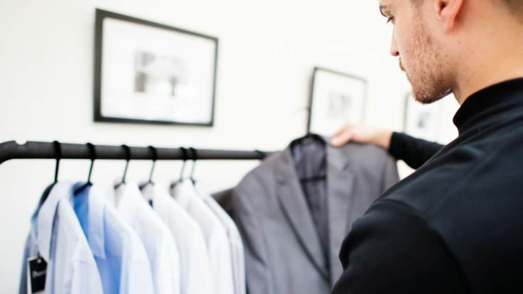 A man in a black shirt looks at clothes on a rack, with shirts and a jacket visible.